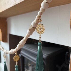 May include: A decorative wooden bead garland with gold filigree charms and teal tassels. The garland features natural wood beads in various shapes and sizes, strung together with a cream-colored cord. The gold charms and teal tassels add a touch of elegance.