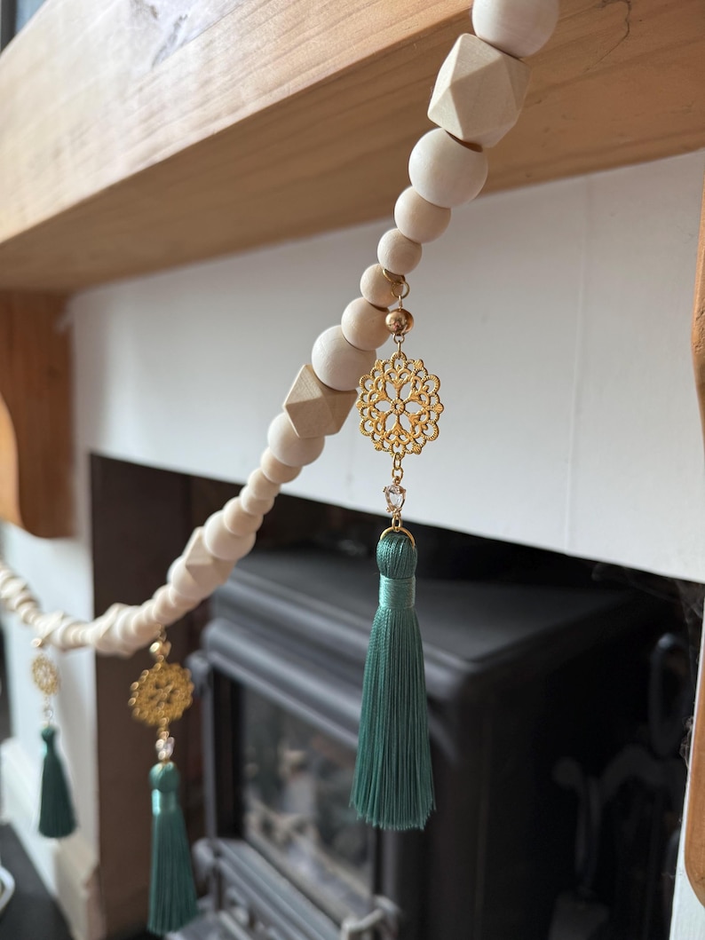 May include: A decorative wooden bead garland with gold filigree charms and teal tassels. The garland features natural wood beads in various shapes and sizes, strung together with a cream-colored cord. The gold charms and teal tassels add a touch of elegance.