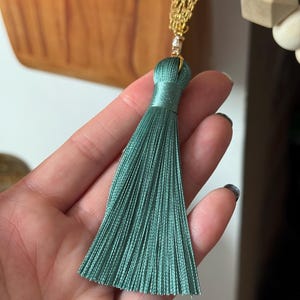 May include: A teal tassel with a gold chain and a small crystal accent. The tassel is made of many strands of teal thread, and the top is wrapped in the same material. The gold chain has a decorative pattern.