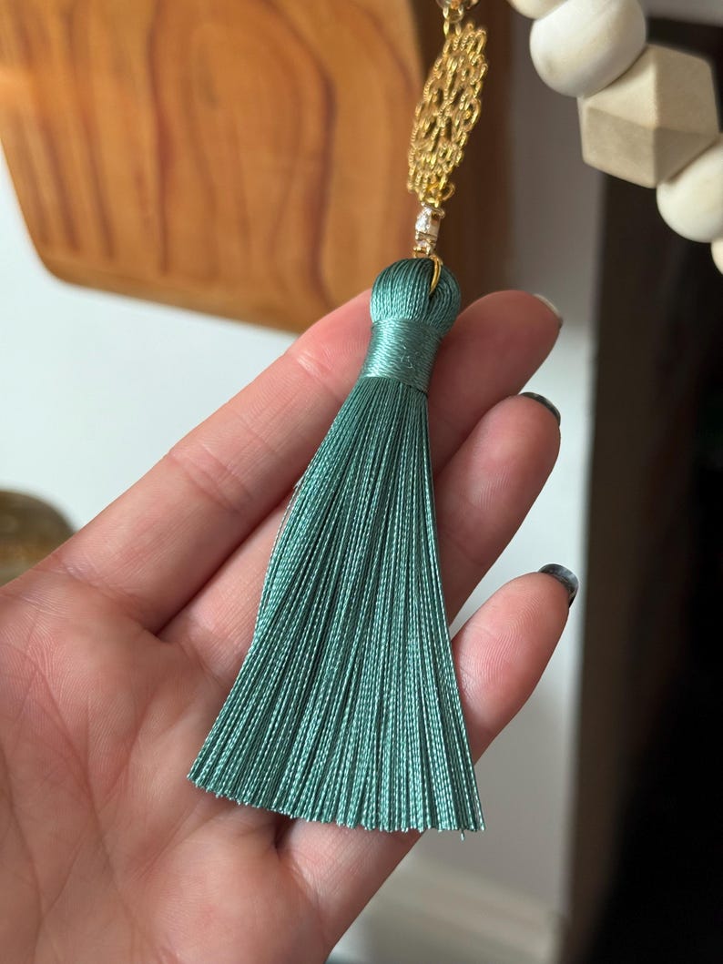 May include: A teal tassel with a gold chain and a small crystal accent. The tassel is made of many strands of teal thread, and the top is wrapped in the same material. The gold chain has a decorative pattern.