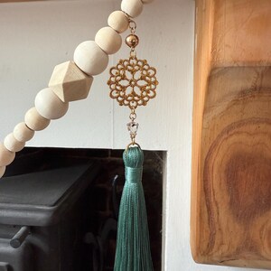 May include: A decorative wooden bead garland with a gold filigree pendant, a clear crystal, and a teal tassel. The garland features natural wood beads and a geometric wooden shape. The pendant is suspended from a gold chain.