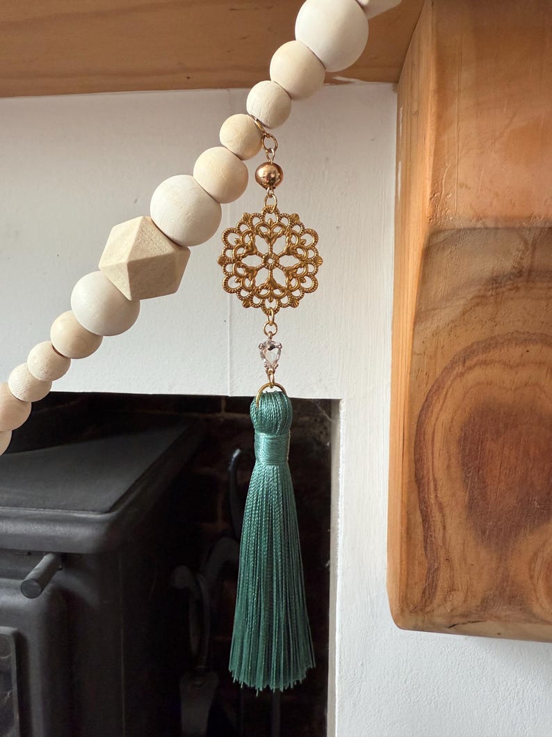 May include: A decorative wooden bead garland with a gold filigree pendant, a clear crystal, and a teal tassel. The garland features natural wood beads and a geometric wooden shape. The pendant is suspended from a gold chain.