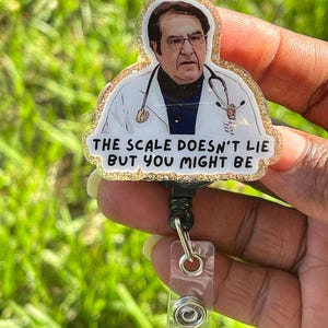 May include: A badge reel featuring a glittery resin topper with an illustration of a doctor wearing a white coat and stethoscope. The text printed on the badge reads, THE SCALE DOESN'T LIE BUT YOU MIGHT BE. The reel has a black plastic housing and a clear strap.