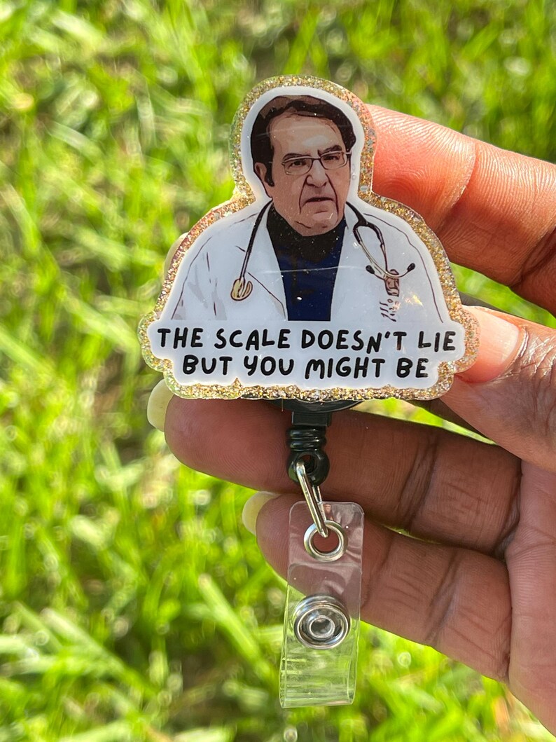 May include: A badge reel featuring a glittery resin topper with an illustration of a doctor wearing a white coat and stethoscope. The text printed on the badge reads, THE SCALE DOESN'T LIE BUT YOU MIGHT BE. The reel has a black plastic housing and a clear strap.