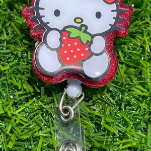 May include: A red glitter badge reel featuring a white cartoon cat holding a red strawberry. The cat has a red bow and black eyes. The badge reel has a clear plastic strap and a metal clip.