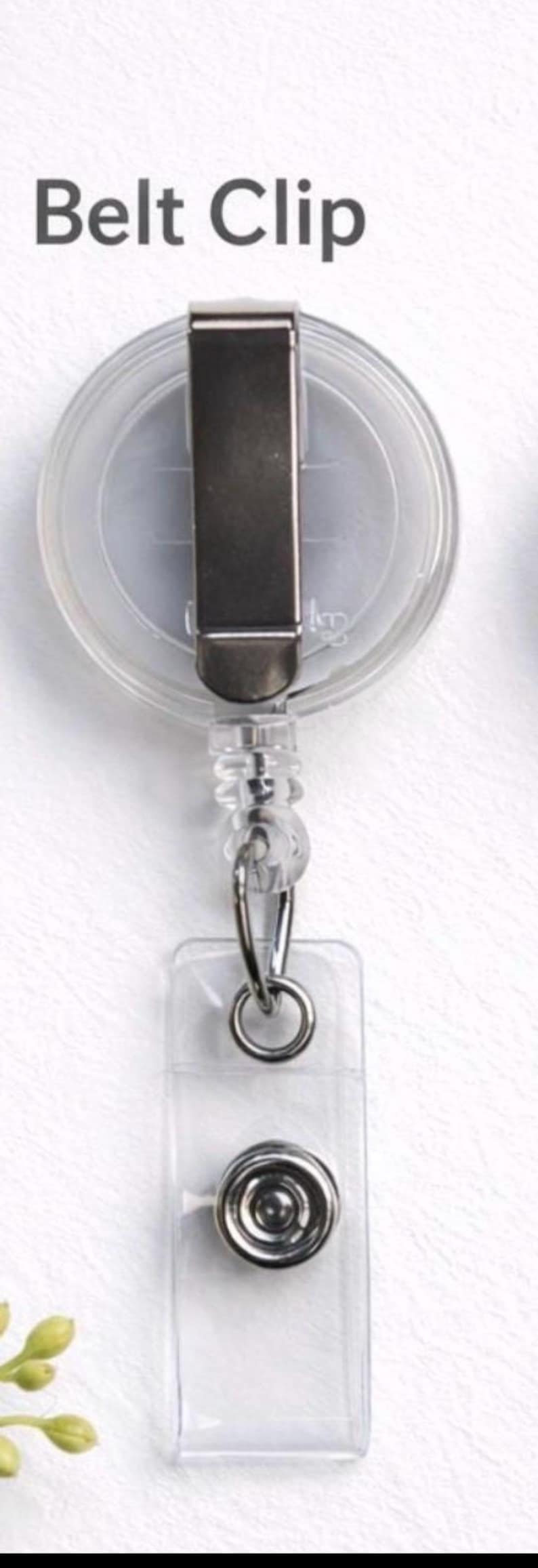 May include: A clear plastic retractable badge reel featuring a metal belt clip on the back and a transparent vinyl strap with a silver-toned metal snap button closure. The text Belt Clip is printed in dark grey above the badge reel.