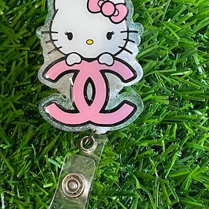 May include: A clear badge reel featuring a white Hello Kitty design with a pink bow and a pink logo. The badge reel has a clear plastic strap and a silver metal snap.