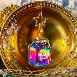 May include: A gold star charm keychain with a rainbow and black holographic charm that says "QUEER Juice" on it. The charm is shaped like a juice carton.