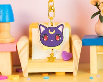 Può includere: Un ciondolo a forma di gatto viola e giallo con una luna crescente e una stella dorata appesa a una catena dorata. Il ciondolo è seduto su una sedia di legno in miniatura con un cuscino bianco.