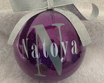 Könnte beinhalten: Lilafarbener Glitzer-Weihnachtsbaumschmuck mit einer silbernen Schleife und dem Namen "Natoya" in weiß geschrieben, mit einem großen silbernen Buchstaben "N" in der Mitte.