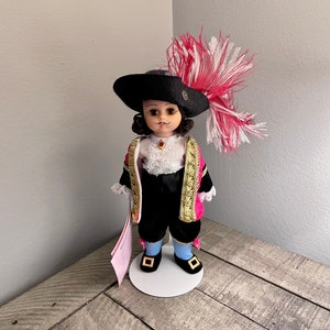 Könnte beinhalten: Eine Porzellanpuppe, die ein schwarzes und pinkfarbenes Kostüm mit einem schwarzen Hut und einer weißen und pinkfarbenen Federboa trägt. Die Puppe steht auf einem weißen Sockel.