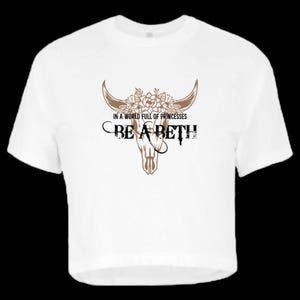May include: A white short-sleeved t-shirt featuring a brown graphic of a cow skull adorned with flowers. The text printed in black reads: IN A WORLD FULL OF PRINCESSES BE A BETH. The design is centered on the chest of the garment.