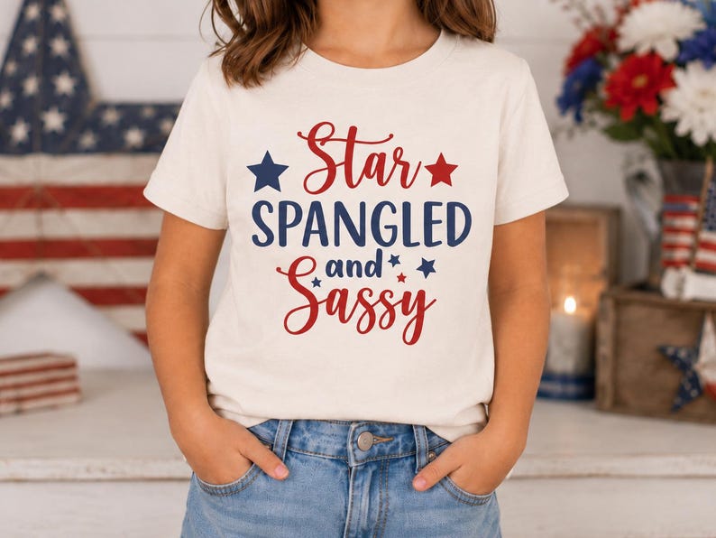 May include: A cream-colored short-sleeve t-shirt featuring the red and blue text Star SPANGLED and Sassy. The design includes small red and blue star graphics surrounding the lettering, which is printed on the front of the cotton garment.