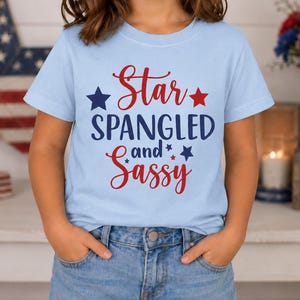 May include: A light blue short-sleeve t-shirt featuring the red and dark blue text Star SPANGLED and Sassy, accented with small dark blue and red stars. The shirt is paired with blue denim jeans, creating a casual look with patriotic colors.