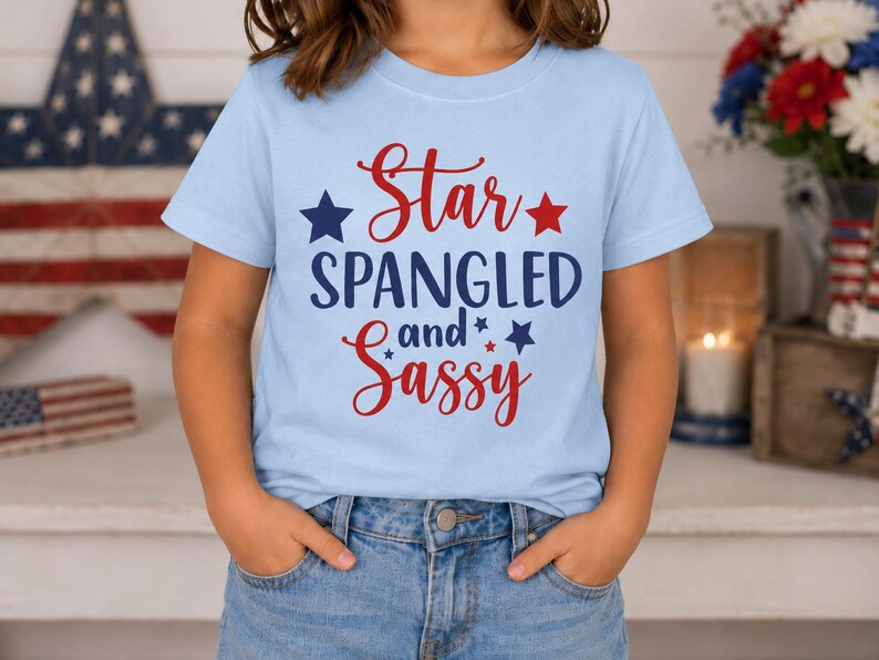 May include: A light blue short-sleeve t-shirt featuring the red and dark blue text Star SPANGLED and Sassy, accented with small dark blue and red stars. The shirt is paired with blue denim jeans, creating a casual look with patriotic colors.