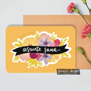 May include: A floral greeting card with the words "asante sana" on a black banner. The card features a bouquet of yellow, purple, and pink flowers against a mustard yellow background. The artist's name, @rafiki.designs, is printed on the card.