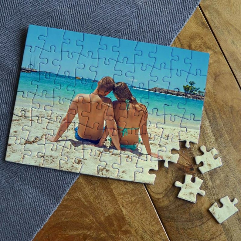 Puede incluir: Un rompecabezas con una imagen de una pareja sentada en una playa. El rompecabezas est&aacute; parcialmente completado, con algunas piezas faltantes.