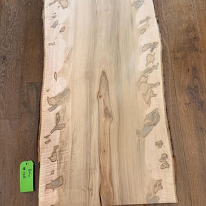 Könnte beinhalten: Eine hellfarbene, rechteckige Holzplatte mit einer lebenden Kante. Die Holzmaserung ist sichtbar, mit dunkleren, unregelmäßigen Mustern. Ein grünes Etikett mit dem Text "Eny #160" ist am Rand befestigt. Das Holz scheint unbehandelt und gebrauchsfertig zu sein.