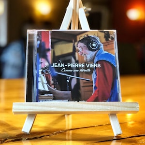 Peut inclure: Un chevalet blanc présentant un CD avec une photo d'un homme portant un casque et une chemise rouge. La pochette du CD indique "JEAN-PIERRE VIENS Comme une eternite EDITION LIMITEE".