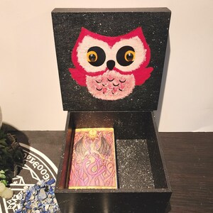 Peut inclure: Une boîte en bois noire avec un couvercle à charnière, ornée d'un hibou rose. La boîte est ouverte, révélant une carte de tarot à l'intérieur. Le fond de la boîte est parsemé d'étoiles.