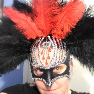 May include: A black mask featuring silver sequin trim and a central dangling red crystal accent. The headpiece is adorned with a large, voluminous arrangement of vibrant red and black ostrich feathers extending outwards from the top.