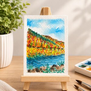 Könnte beinhalten: Ein kleines Aquarellbild auf einer Holzstaffelei zeigt eine lebendige Herbstlandschaft mit blauem Himmel, einem mit orangefarbenen, gelben und roten Bäumen bewachsenen Hügel und einem blauen See im Vordergrund mit grünem Gras und braunen Felsen.
