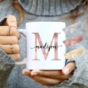 May include: White ceramic mug with a rose gold letter "M" and the name "madison" in cursive script. The mug is being held by a person wearing a gray sweater.