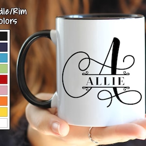 May include: White ceramic mug with a black rim and handle. The mug features a black cursive letter "A" with a decorative flourish and the name "ALLIE" written in black below the letter. The mug is being held by a person's hand.