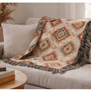 May include: A woven throw blanket with a geometric pattern in orange, blue, and cream tones, draped over a beige sofa. The blanket has a fringed edge. A coffee table with books and a mug is in the foreground.