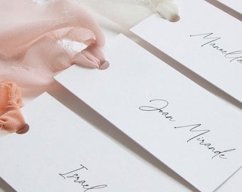 May include: Rectangular white paper place cards with rounded corners and a hole punched at the top. Each card features black cursive text with names such as Joan Mirande and Mannella Barragan. Soft pink and white frayed silk ribbons are tied through the holes.