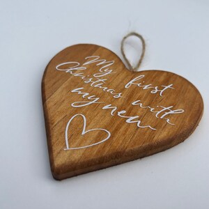 May include: Wooden heart-shaped Christmas ornament with the text "My first Christmas with my new" in white script. A small white heart is drawn below the text. The ornament has a natural wood grain and a jute string for hanging.