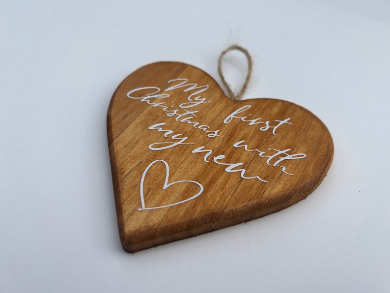 May include: Wooden heart-shaped Christmas ornament with the text "My first Christmas with my new" in white script. A small white heart is drawn below the text. The ornament has a natural wood grain and a jute string for hanging.