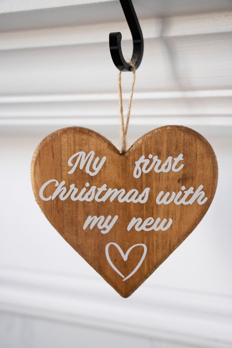 May include: Wooden heart-shaped Christmas ornament with the text "My first Christmas with my new" in white script. A white heart is drawn below the text. The ornament hangs from a jute string.