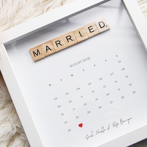 May include: A white square shadow box frame containing a calendar for August 2021. The word MARRIED is spelled out in wooden letter tiles at the top. A small red heart marks the 24th, and the names Sarah Neville & Rafa Barragan are printed below.