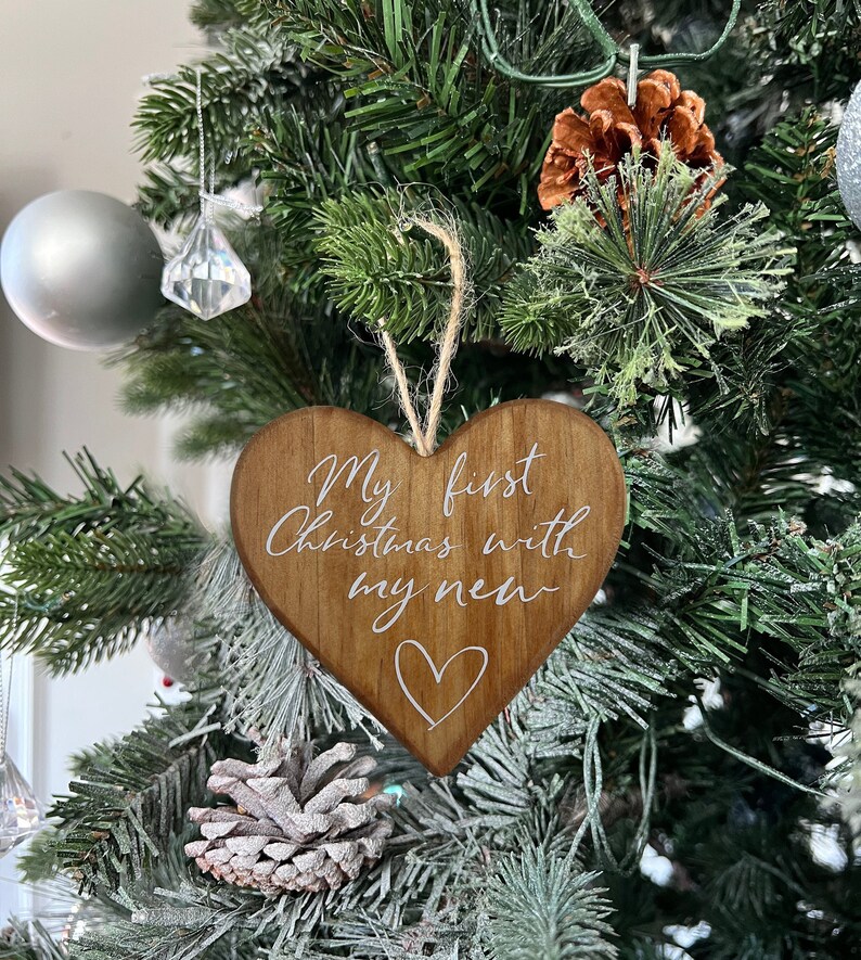 May include: A wooden heart-shaped Christmas ornament with the text "My first Christmas with my new" in white script. A white heart is drawn below the text. The ornament hangs from a natural twine string. The ornament is hanging on a Christmas tree.