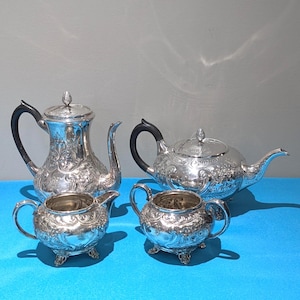 May include: A silver tea set with detailed floral patterns. The set comprises a coffee pot, a teapot, a creamer, and a sugar bowl. The coffee pot and teapot have black handles. The set is displayed on a blue surface.
