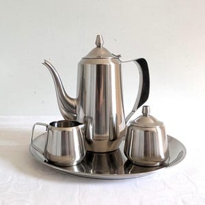 May include: A stainless steel coffee or tea service set. The set includes a tall coffee pot, a creamer, a sugar bowl with a lid, and a serving tray. The items are all made of shiny, reflective metal.