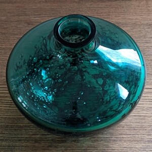 Puede incluir: Un jarr&oacute;n de vidrio redondo de color verde azulado, con un cuerpo ancho y bulboso y un cuello estrecho. El vidrio tiene una apariencia texturizada con peque&ntilde;as burbujas, lo que le da un aspecto artesanal &uacute;nico. El jarr&oacute;n est&aacute; sobre una superficie de madera.