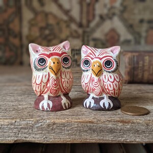 May include: Two small owl figurines featuring hand-painted red and white patterns on a cream base. Each owl has a yellow beak, large black eyes, and white talons resting on a dark brown base. A copper penny is placed next to the smaller owl.