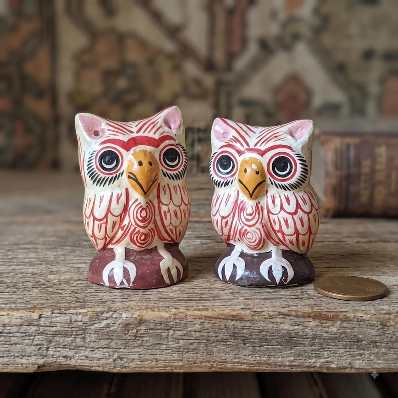 May include: Two small owl figurines featuring hand-painted red and white patterns on a cream base. Each owl has a yellow beak, large black eyes, and white talons resting on a dark brown base. A copper penny is placed next to the smaller owl.