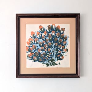 May include: Framed art print featuring a peacock design. The artwork showcases a peacock with a fan of orange, blue, and green brushstrokes. The print is set against a light peach mat and enclosed in a dark brown wooden frame.