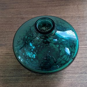 Puede incluir: Un jarr&oacute;n redondo de vidrio color verde azulado, con una base ancha y plana y una abertura estrecha. El vidrio tiene una apariencia texturizada y burbujeante, y refleja la luz. El jarr&oacute;n est&aacute; sobre una superficie de madera. El di&aacute;metro del jarr&oacute;n es de unos 20 cm.