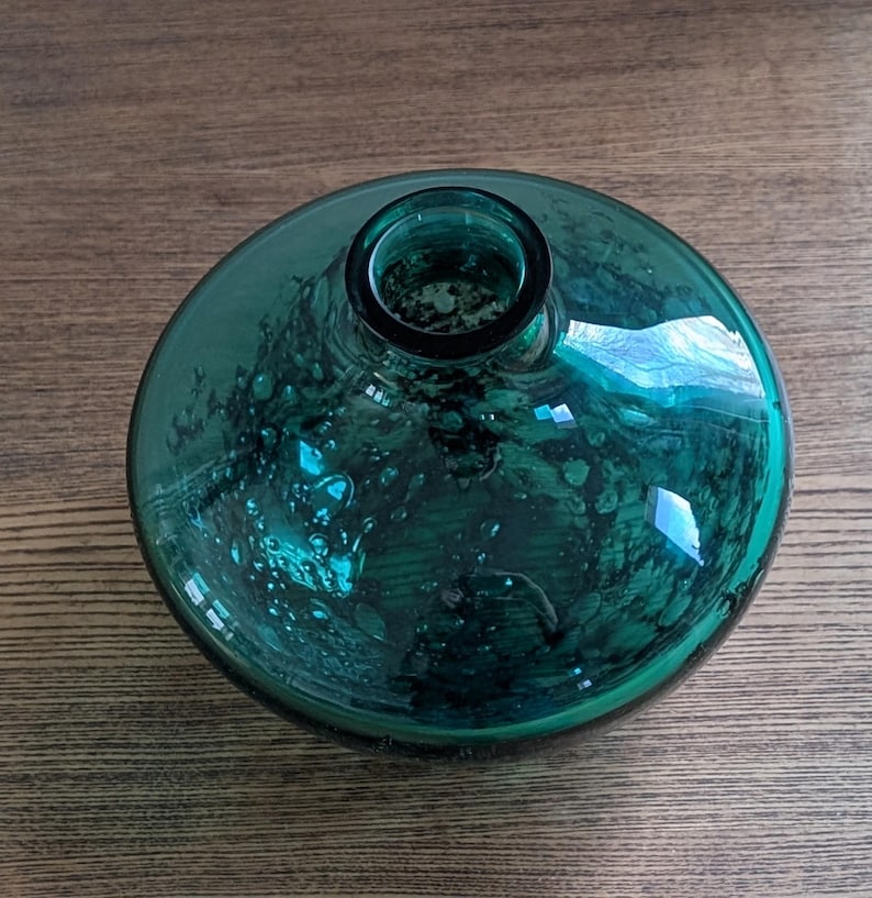Puede incluir: Un jarr&oacute;n redondo de vidrio color verde azulado, con una base ancha y plana y una abertura estrecha. El vidrio tiene una apariencia texturizada y burbujeante, y refleja la luz. El jarr&oacute;n est&aacute; sobre una superficie de madera. El di&aacute;metro del jarr&oacute;n es de unos 20 cm.
