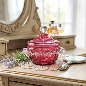 May include: A deep pink glass candy dish with a matching lid featuring a clear glass finial knob. The bowl has a textured, ribbed body and a decorative clear glass ruffled edge trim around the centre. The glass is translucent and vibrant.
