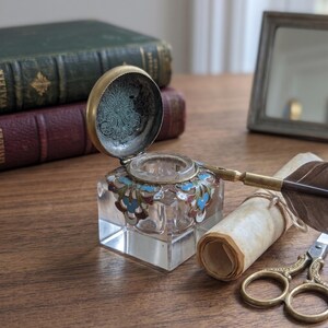 May include: A clear glass inkwell with a hinged brass lid featuring intricate floral enamel detailing in blue and red. The inkwell is accompanied by a quill pen with a dark wooden handle and a pair of ornate brass scissors resting near a rolled parchment.