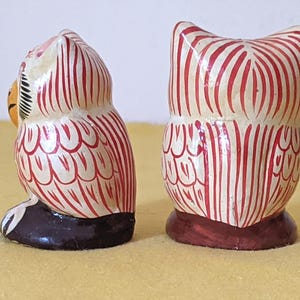 May include: Two small owl figurines featuring a cream-colored base with hand-painted red vertical stripes and feather patterns. One owl is shown from the side, revealing a yellow beak and black base, while the other is shown from the back.