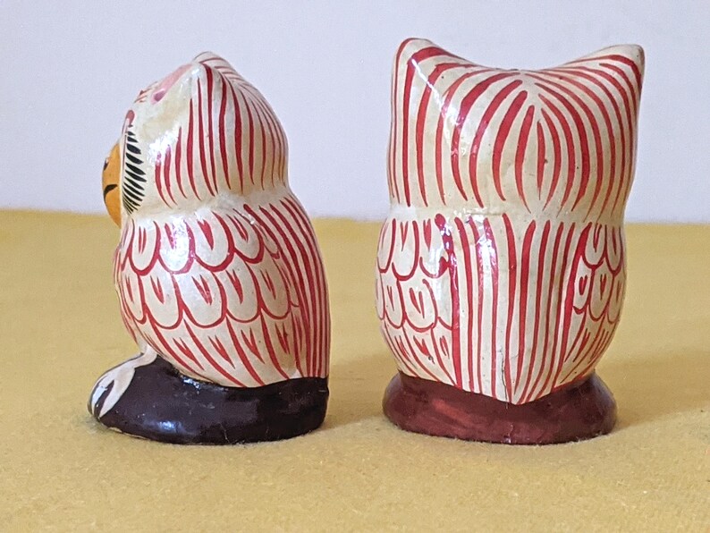 May include: Two small owl figurines featuring a cream-colored base with hand-painted red vertical stripes and feather patterns. One owl is shown from the side, revealing a yellow beak and black base, while the other is shown from the back.