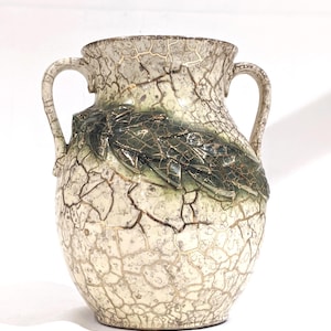 May include: A ceramic vase featuring a cream-coloured base with a dark brown crackle glaze pattern. The vase has two side handles and a prominent, raised relief decoration of dark green leaves wrapped around the upper body of the vessel.