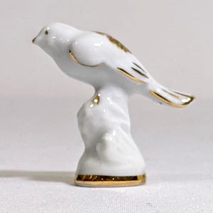 May include: A white porcelain bird figurine perched on a textured base. The bird features delicate gold trim along its wings, tail, and the base of the stand. The surface is smooth and glossy, with a small brown accent visible on the bird's back.