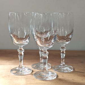 May include: Four clear, stemmed wine glasses. The glasses have a classic design with a rounded bowl and a decorative stem. The bases are round and the glasses are arranged on a wooden surface.